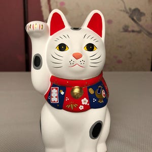 May include: A white ceramic Maneki Neko cat figurine with red ears, a red and blue collar, and a gold bell. The cat has black markings and is in a beckoning pose, with one paw raised. The figurine is a traditional Japanese symbol of good luck and fortune.