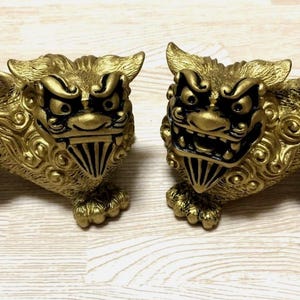 May include: Two golden lion statues with detailed features, including expressive faces and ornate manes. The statues are posed side-by-side, showcasing their intricate design and craftsmanship. The statues are on a light-colored surface.