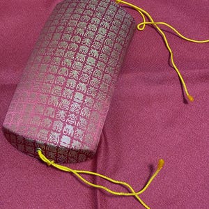 Puede incluir: Una bolsa cilíndrica de tela rosa con un patrón dorado repetido. La bolsa tiene un cierre de cordón amarillo. El fondo es una tela rosa lisa.