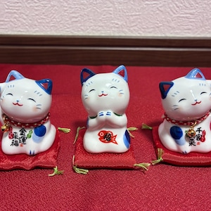 Op de afbeelding: Drie witte keramische Maneki Neko beeldjes met blauwe accenten, rode halsbanden en gouden belletjes. Elke kat zit op een klein rood kussen. Eén kat heft een poot, een andere vouwt zijn poten en de derde heft een poot.