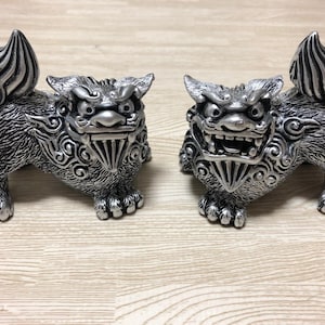 May include: A pair of silver-colored guardian lion statues, known as Komainu, featuring intricate fur textures, swirling patterns on the shoulders, and fierce facial expressions with bared teeth. Each figure stands in a dynamic pose with a raised tail.