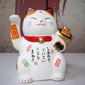 Może przedstawiać: Biała ceramiczna figurka Maneki-neko z uniesioną prawą łapą. Kot ma czarne wąsy, czerwony kołnierz i złotą torbę na monety. Japoński tekst jest widoczny na ciele i ramieniu. Uszy i głowa kota są zaakcentowane czernią i złotem.