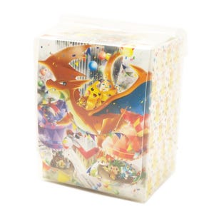 May include: A clear plastic box with a vibrant illustration of Charizard and Pikachu from the Pokémon series. The box is filled with small, confetti-like shapes and is sealed with a clear plastic lid. The box is square-shaped.