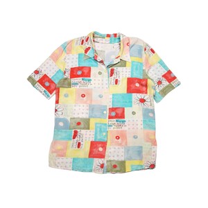 Puede incluir: Camisa de manga corta con diseño de patchwork. La camisa presenta un collage de cuadrados coloridos en rojo, amarillo, azul y verde, con detalles de margaritas y texto. La camisa tiene cuello y está hecha de tela ligera.