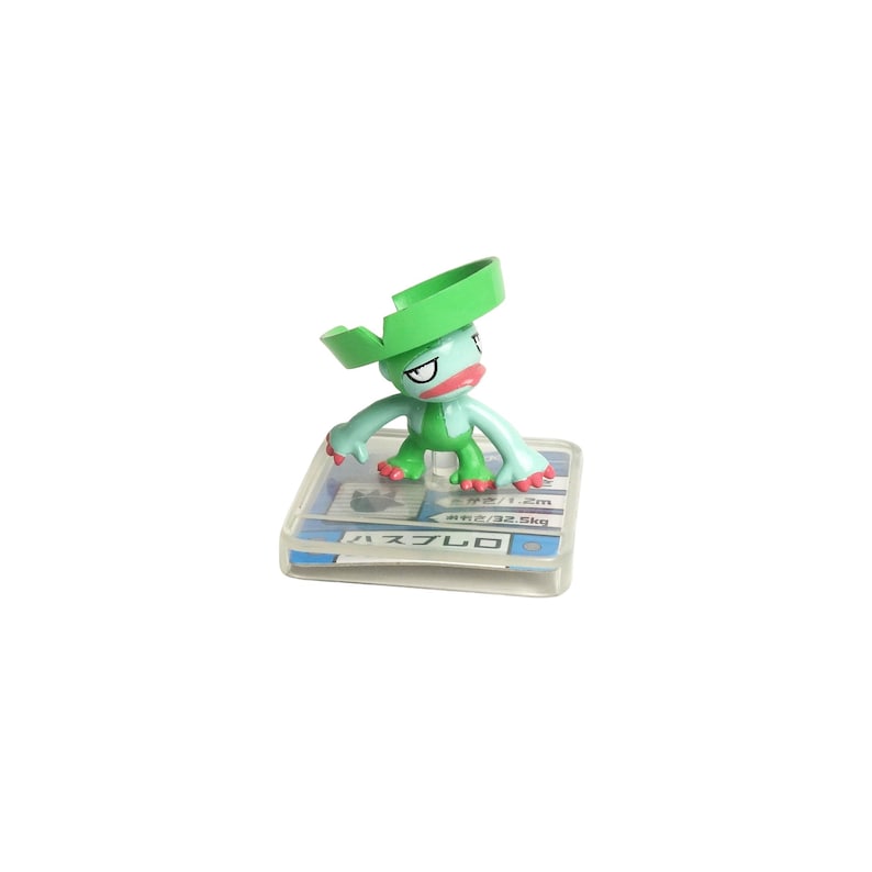 May include: A green and blue figurine of a cartoon character with a large green hat. The figurine is standing on a clear plastic base with Japanese text. The character has a red mouth and is in a playful pose.