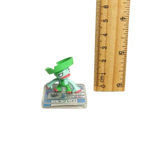May include: A small, green figurine of a cartoon character with a green hat, red mouth, and blue arms and legs. The figurine is on a clear base with Japanese text and measurements. A wooden ruler is next to the figurine.
