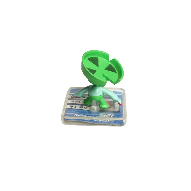 May include: A green and light blue figurine of a character with a leaf-shaped head, standing on a clear plastic base. The figurine has outstretched arms and red accents on its feet and hands. The base has text in a foreign language.