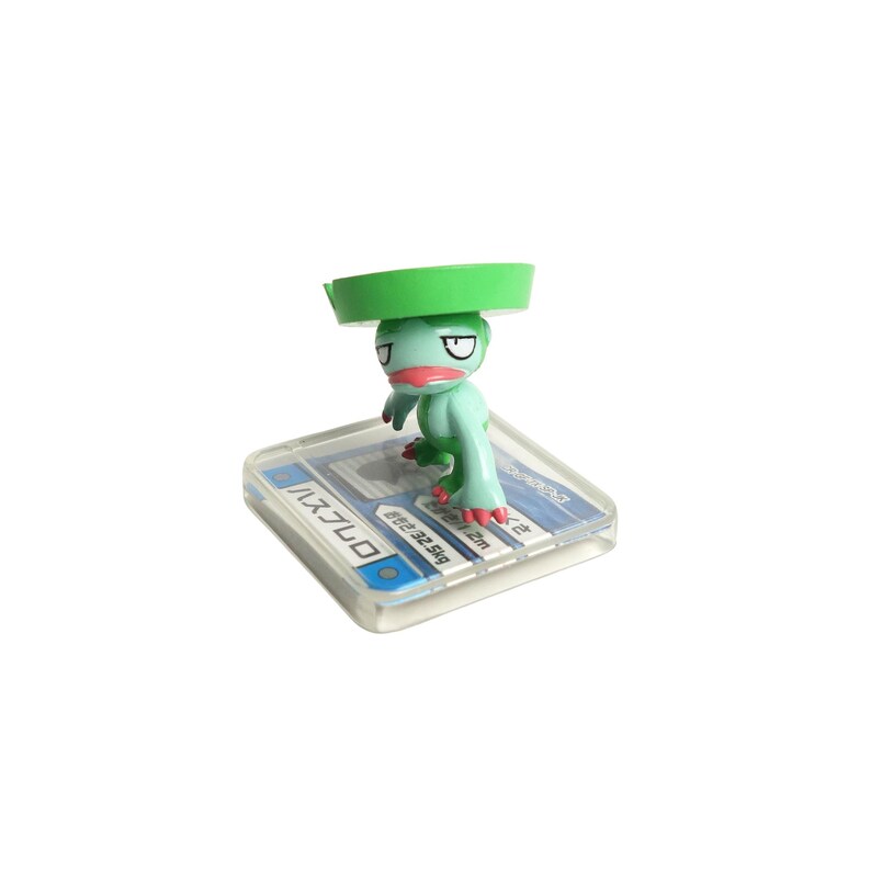 May include: A small, light green figurine of a frog-like creature with a green hat. The figurine has red accents on its feet and mouth. It stands on a clear, rectangular base with Japanese text.