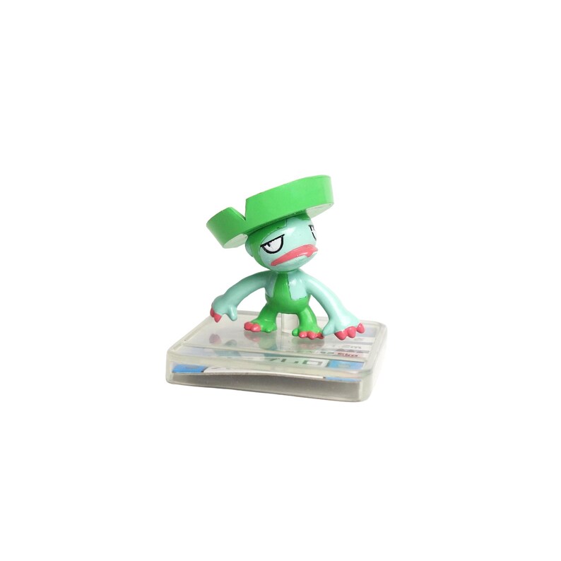 May include: A small, green cartoon figure with a large, flat, green hat and red lips. The figure is standing on a clear, rectangular base. The figure has a grumpy expression and is a collectible item.