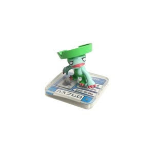 May include: A small, green and red figurine of a cartoon character with a large green headpiece. The figure is on a clear plastic base with Japanese text and blue accents. The figurine is in a crouching pose.
