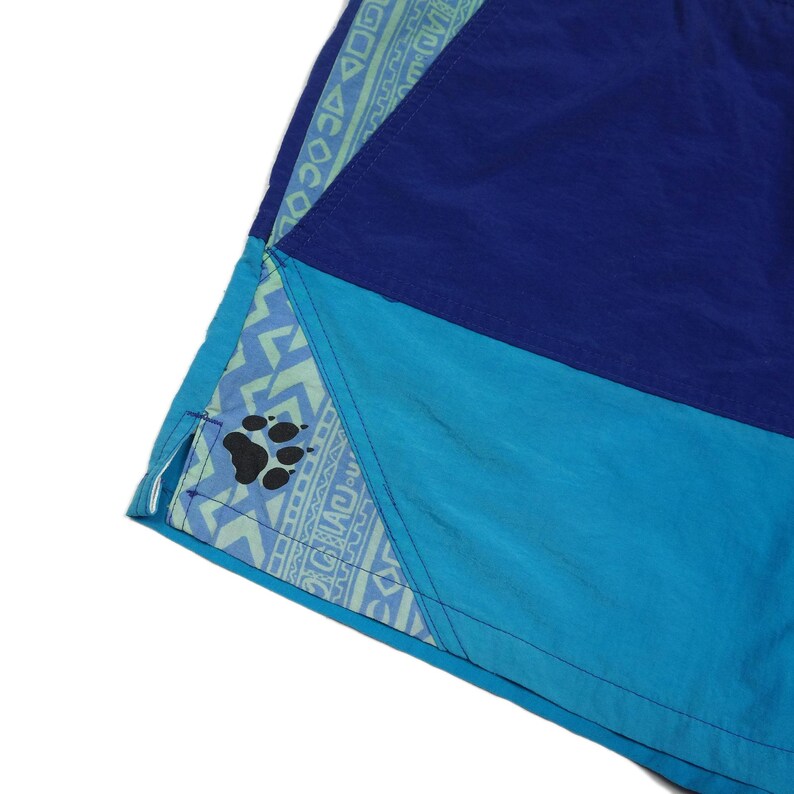 Puede incluir: Primer plano de la parte inferior de un par de pantalones cortos azules y turquesa con un ribete estampado en verde y azul. El ribete presenta una huella de pata.