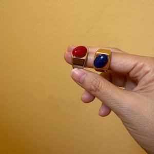 May include: Two gold-toned rings featuring oval-shaped cabochon stones. One ring displays a smooth, vibrant red stone, while the other ring features a deep blue stone with subtle natural variations. Both rings have a wide, polished metal band design.