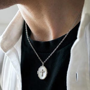 May include: Silver-toned necklace with a cross pendant. The pendant has an oval shape with a mother-of-pearl background and a detailed cross design. The necklace is worn over a black shirt and a white collared shirt.