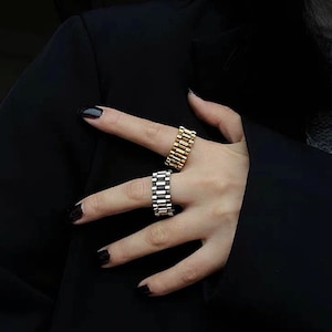 May include: Two rings, one gold-toned and one silver-toned, are displayed on a hand. The rings have a watch band style design. The background is a dark, solid color, which makes the rings stand out.