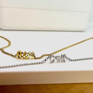 May include: Two gold and silver necklaces with personalized charms. The gold necklace has the letters "B&N" and the silver necklace has the letters "C&K" with a heart in between.
