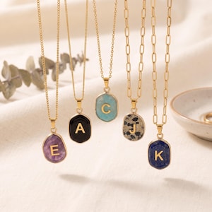 May include: Five gold-toned necklaces featuring irregular, stone-like pendants in purple, black, light blue, spotted white, and dark blue. Each pendant displays a gold-colored letter: E, A, C, J, and K. The necklaces hang from delicate gold chains.