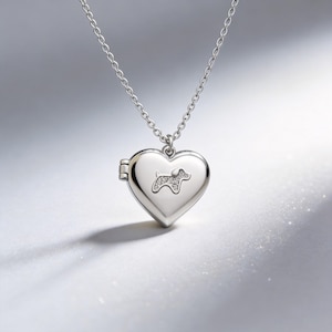 May include: A silver heart-shaped locket necklace with a small dog silhouette embellished with clear crystals. The locket hangs from a delicate chain. The locket is closed, and the background is a soft, neutral tone.