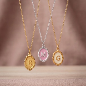 May include: Three oval pendant necklaces featuring scalloped edges. The left pendant is gold with a letter B, the centre is silver with a pink enamel background and letter M, and the right is gold with a cream enamel background and letter G.