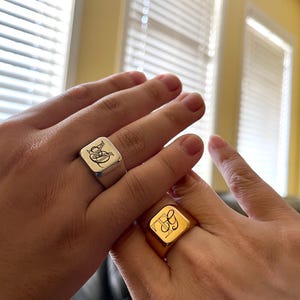 May include: Two square signet rings with engraved initials. The ring on the left is silver and the ring on the right is gold.