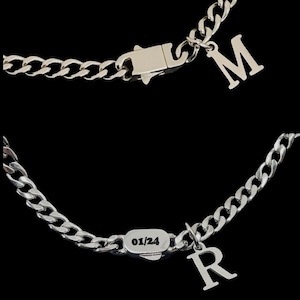 May include: Silver curb chain necklace with a silver charm pendant. The charm is a letter 'M' on one side and '01/24' on the other side. The necklace also has a silver clasp.