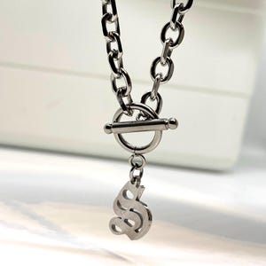 May include: A silver chain necklace with a toggle clasp and a silver dollar sign pendant.