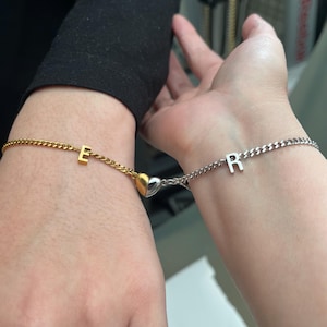 May include: Two matching bracelets with a heart charm connecting them. One bracelet is gold with the letter "E" and the other is silver with the letter "R".