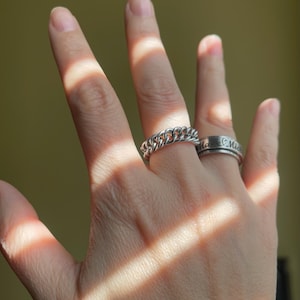 May include: A hand wearing two silver rings. The ring on the index finger is a chain link style ring. The ring on the middle finger is a thin band with the word "Mia" engraved on it.