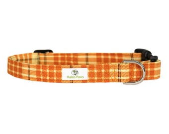 May include: An orange and cream plaid dog collar with a black buckle and a silver metal D-ring. The collar has a white label with the text "Hala's Paws".