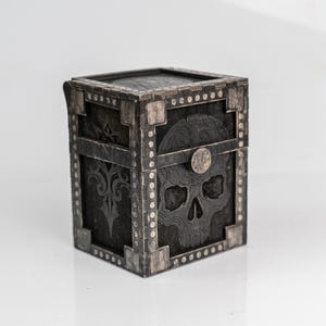 Könnte beinhalten: Eine quadratische, dunkelgraue Box mit einem Totenkopf-Design auf der Vorderseite. Die Box hat silberfarbene Metallakzente, darunter Eckstücke und dekorative Nieten. Der Deckel ist ebenfalls dunkelgrau und strukturiert.