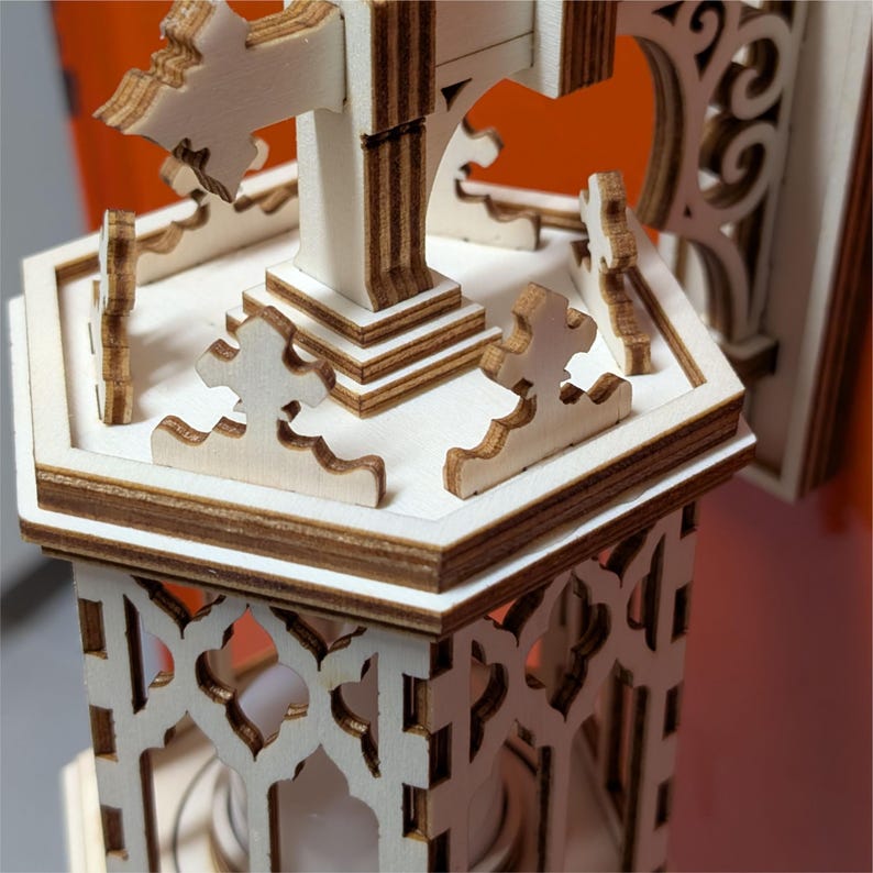 May include: A detailed wooden model of a church or cathedral, featuring intricate cut-out designs and layered construction. The model has a light wood color with darker brown edges, showcasing the architectural details. The design includes a cross and decorative elements.