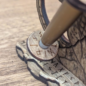 May include: Close-up of a wooden desk accessory with engraved runes. The accessory features a circular piece with runes and a brown pen holder. The wood has a natural grain and a warm, brown tone. The design appears to be laser-cut.
