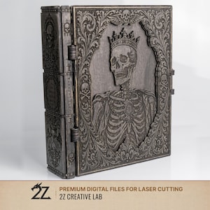 Pode incluir: Uma caixa de madeira cinza escuro com uma porta articulada, apresentando um design esculpido detalhado de um esqueleto coroado dentro de uma moldura ornamentada. A caixa tem uma construção em camadas com elementos esculpidos e texto adicionais. A parte inferior da imagem inclui o texto "PREMIUM DIGITAL FILES FOR LASER CUTTING" e "2Z CREATIVE LAB".