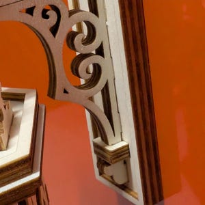 May include: Close-up of a wooden architectural detail with intricate scrollwork and layered construction. The design features a light brown color with darker brown accents, set against an orange background. The piece appears to be part of a larger structure.