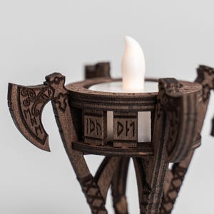 Pu&ograve; includere: Un portacandele in legno con un design decorativo a forma di ascia. Il supporto marrone ha una parte superiore circolare con una candela LED bianca. Il design include rune scolpite e motivi geometrici. Il portacandele &egrave; progettato per una piccola candela.