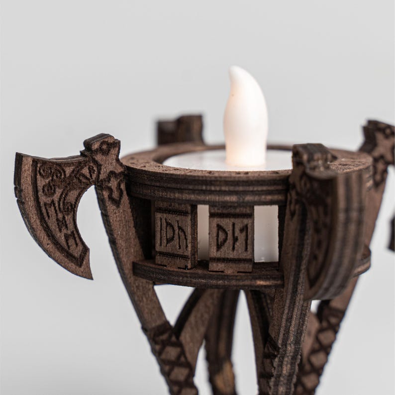 Pu&ograve; includere: Un portacandele in legno con un design decorativo a forma di ascia. Il supporto marrone ha una parte superiore circolare con una candela LED bianca. Il design include rune scolpite e motivi geometrici. Il portacandele &egrave; progettato per una piccola candela.