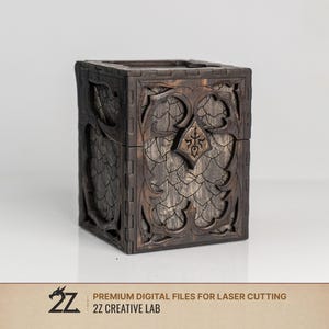 Könnte beinhalten: Eine dunkelbraune Holzbox in quadratischer Form mit Deckel. Die Außenseite zeigt aufwendige Laserschnittmuster, die an Drachenschuppen und verzierte gotische Bögen erinnern. Ein zentrales schildförmiges Emblem befindet sich auf der Vorderseite.