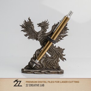 May include: A wooden pen holder featuring a detailed phoenix design. The pen holder is brown and holds a gold pen. The base of the holder has the text "PREMIUM DIGITAL FILES FOR LASER CUTTING" and "2Z CREATIVE LAB".