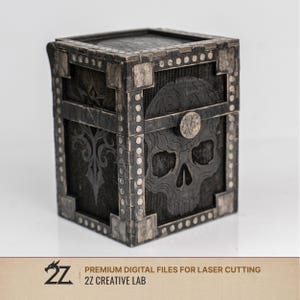 Può includere: Una scatola di legno grigio scuro tagliata al laser con un disegno a teschio sul davanti. La scatola ha dettagli metallici decorativi e un coperchio incernierato. Il testo "PREMIUM DIGITAL FILES FOR LASER CUTTING" e "2Z CREATIVE LAB" è in basso.