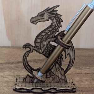 May include: A wooden dragon-shaped pen holder with a gold pen inserted. The dragon is intricately carved with detailed scales and wings. The base of the holder has etched symbols. The pen is a metallic gold color.