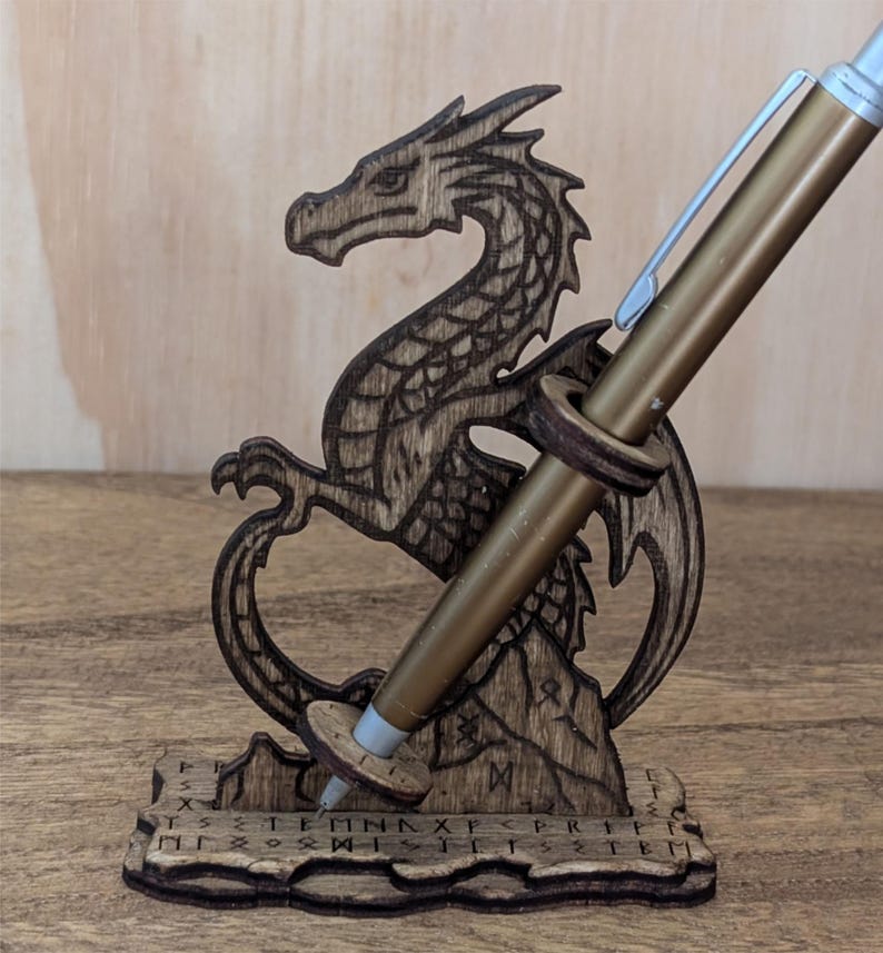 May include: A wooden dragon-shaped pen holder with a gold pen inserted. The dragon is intricately carved with detailed scales and wings. The base of the holder has etched symbols. The pen is a metallic gold color.