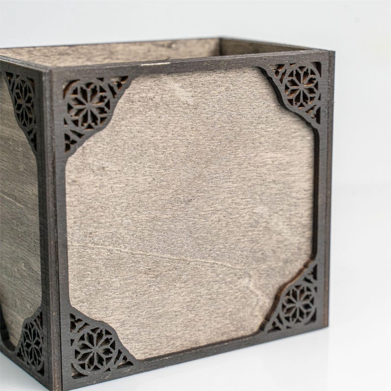 May include: A square wooden box with a light gray interior and dark brown, laser-cut decorative corners. The box has a simple, geometric design, suitable for storage or display.