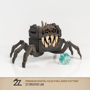 May include: A dark brown, multi-legged creature-shaped box with large, pointed, off-white teeth and red accents. The box features intricate carvings and is positioned next to a translucent, teal-colored twenty-sided die. Text reads 2Z CREATIVE LAB.
