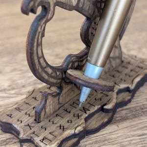 May include: A wooden pen holder with a gold-colored pen. The holder is intricately carved with a Viking ship design and runes. The pen rests in a small, round wooden support. The wood has a dark brown stain.