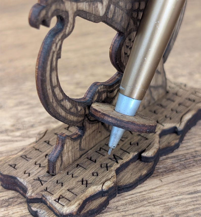 May include: A wooden pen holder with a gold-colored pen. The holder is intricately carved with a Viking ship design and runes. The pen rests in a small, round wooden support. The wood has a dark brown stain.