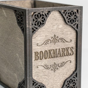 May include: A wooden box with the word "BOOKMARKS" engraved on the front. The box has a light gray wood grain finish with dark gray decorative trim and corner accents. The design is a decorative storage container.