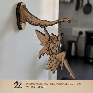 May include: A wooden fairy sculpture hanging from a branch, mounted on a wall. The fairy has detailed wings and is swinging from a rope. The branch is a light brown colour, and the fairy is a lighter shade of brown. The text "2Z CREATIVE LAB" is at the bottom.