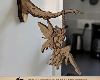 Może przedstawiać: Drewniana rzeźba wróżki wiszącej na gałęzi, przymocowanej do ściany. Wróżka ma szczegółowe skrzydła i huśta się na linie. Gałąź jest w jasnobrązowym kolorze, a wróżka jest w jaśniejszym odcieniu brązu. Tekst "2Z CREATIVE LAB" znajduje się na dole.