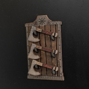 Może przedstawiać: Prostokątna drewniana tablica ścienna z trzema miniaturowymi toporami zamontowanymi poziomo. Tablica posiada wyrzeźbiony symbol Vegvisir u g&oacute;ry oraz rząd znak&oacute;w runicznych wzdłuż dolnej krawędzi. Każdy top&oacute;r ma ciemny trzonek i ostrze.