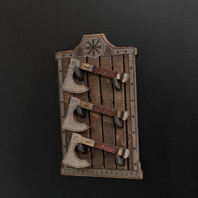Może przedstawiać: Prostokątna drewniana tablica ścienna z trzema miniaturowymi toporami zamontowanymi poziomo. Tablica posiada wyrzeźbiony symbol Vegvisir u g&oacute;ry oraz rząd znak&oacute;w runicznych wzdłuż dolnej krawędzi. Każdy top&oacute;r ma ciemny trzonek i ostrze.