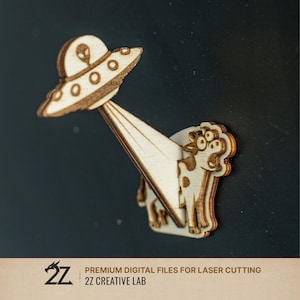 Può includere: Un design in legno tagliato al laser raffigurante un UFO che emette un raggio su una mucca. L'UFO ha una testa aliena e la mucca ha macchie e un'espressione sorpresa. Il design è realizzato in legno marrone chiaro.
